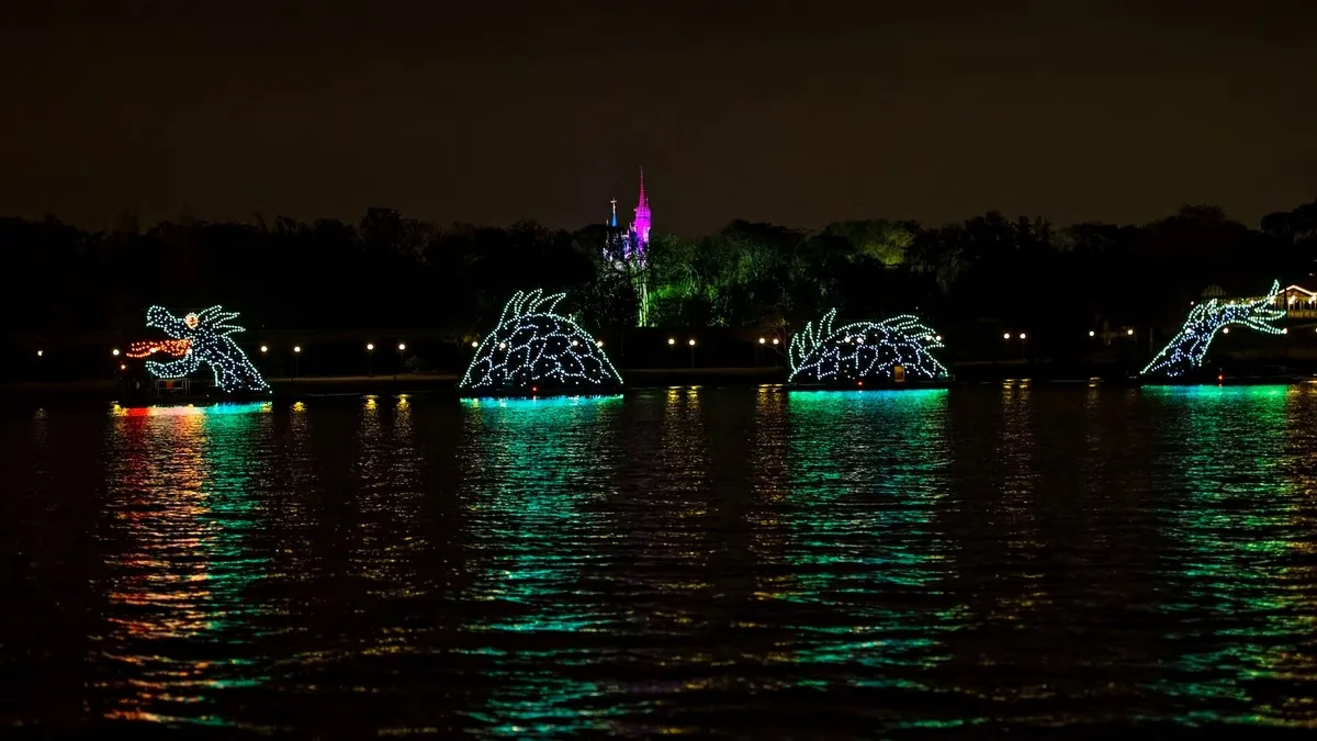 An illuminated dragon float glides across a dark, reflective lake, with green and blue lights outlining their forms. In the background, a softly lit castle stands against the night sky.