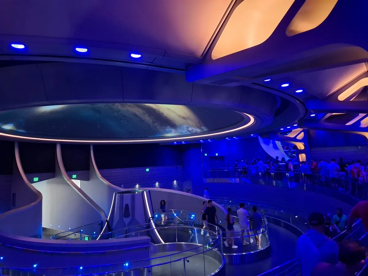 A futuristic indoor space features a curved walkway filled with people, illuminated by blue lighting. The ceiling displays an image of space, adding to the sci-fi atmosphere.