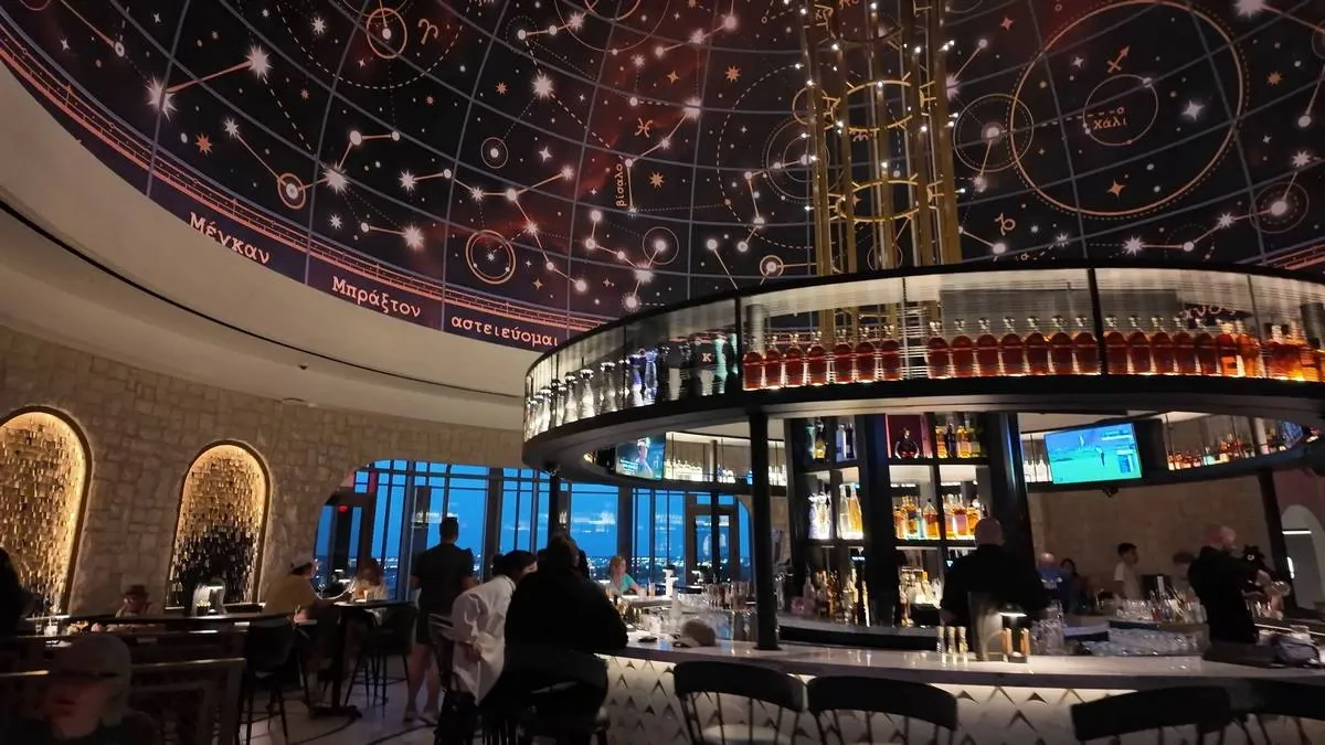 A stylish bar with a circular counter is bustling with patrons, beneath a dome ceiling adorned with a celestial map. Bottles of liquor are displayed on shelves around the bar, and a large screen shows a sports match in the background.