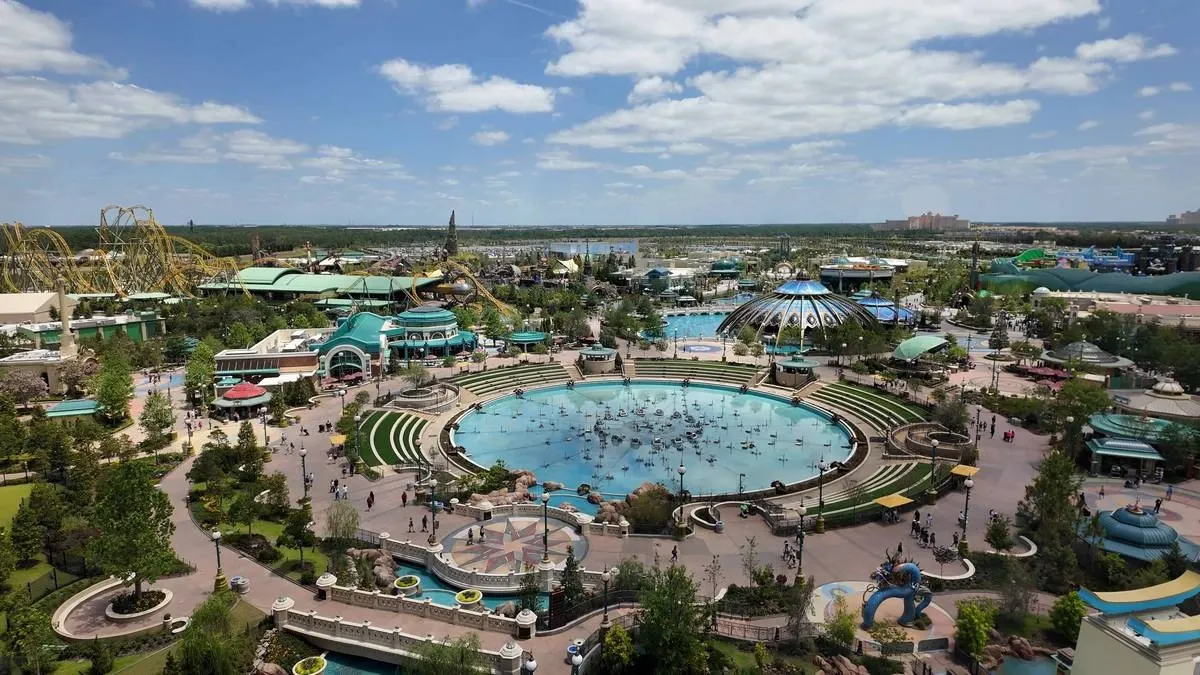 Aerial view of an amusement park featuring a large circular water attraction at the center, surrounded by greenery and winding pathways. Roller coasters and various themed buildings are visible in the background under a partly cloudy sky.