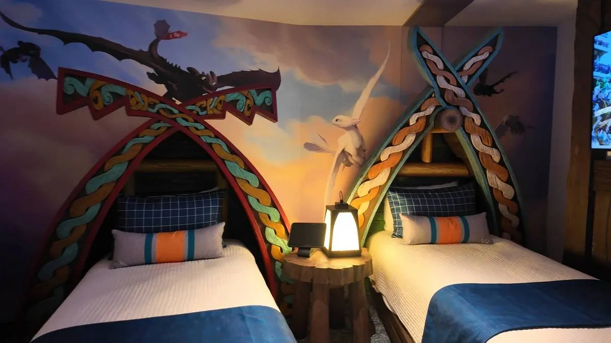 A themed bedroom featuring two twin beds with colorful, Viking-inspired headboards and bedding in blue and white. A mural of flying dragons adorns the wall above, and a rustic wooden nightstand with a lantern sits between the beds.
