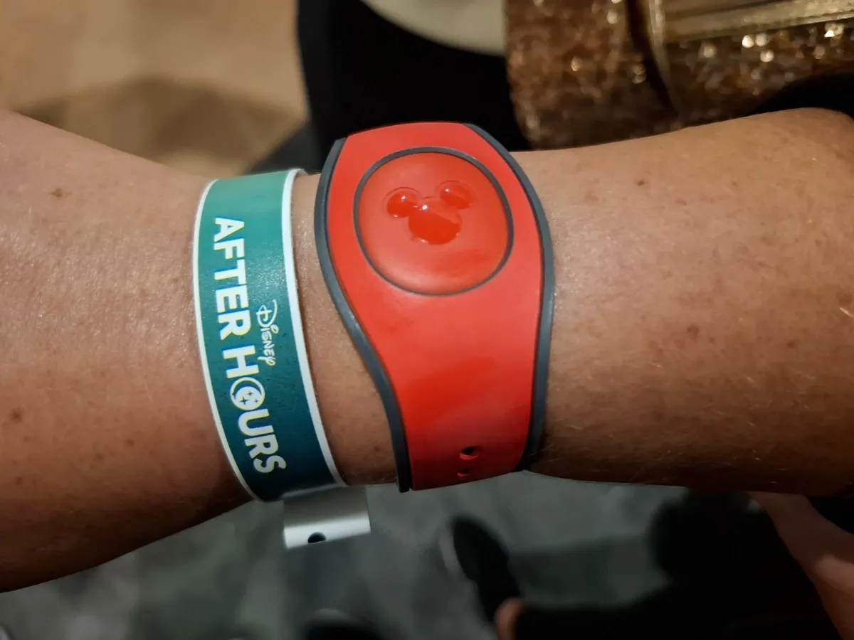 A wrist wearing a red Disney MagicBand with a Mickey Mouse logo, alongside a green wristband labeled "Disney After Hours.".