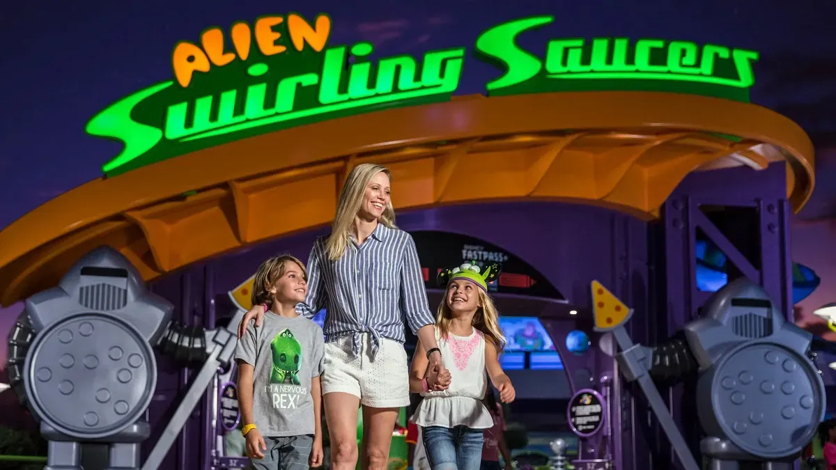 A smiling woman and two children walk happily in front of the "Alien Swirling Saucers" ride entrance, featuring a vibrant green and orange sign. The children wear playful alien-themed hats, and the setting is lively and colorful.