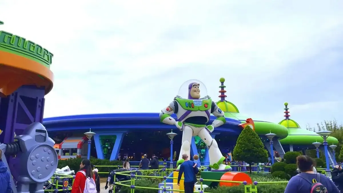 A giant Buzz Lightyear statue stands prominently in a colorful amusement park setting, surrounded by vibrant green and blue structures. Visitors are walking around, enjoying the lively atmosphere under a partly cloudy sky.