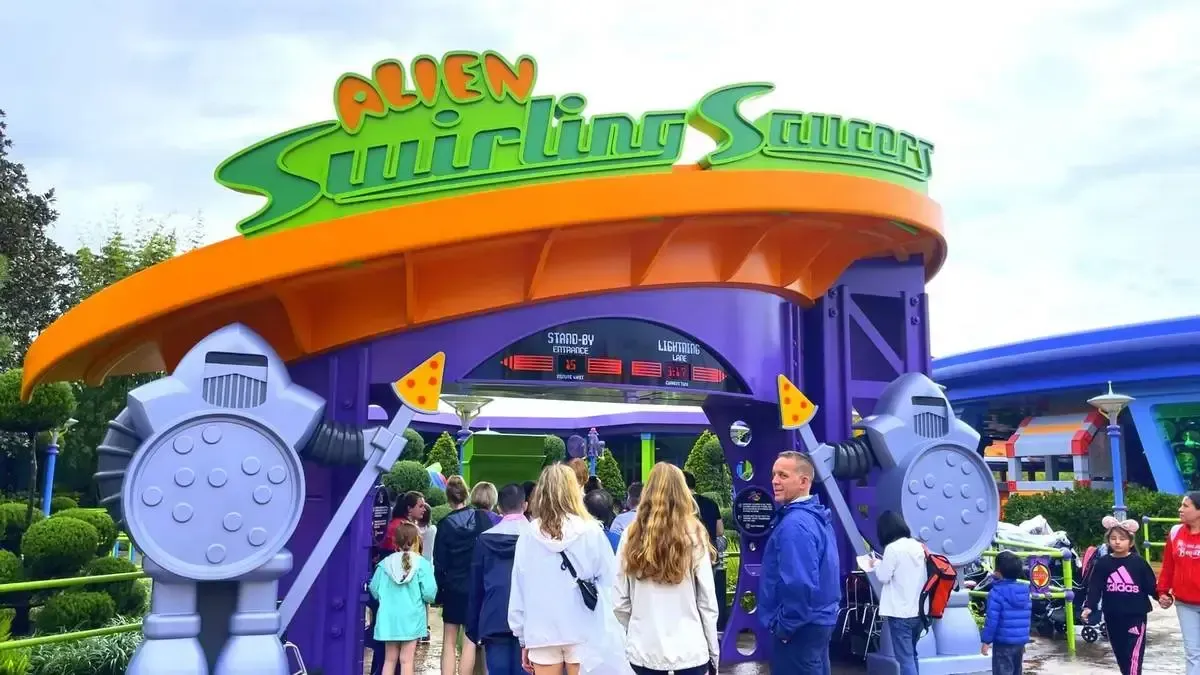 Visitors gather at the entrance of the "Alien Swirling Saucers" ride, featuring a sign with green and orange colors. The entrance is flanked by whimsical alien-themed decorations.
