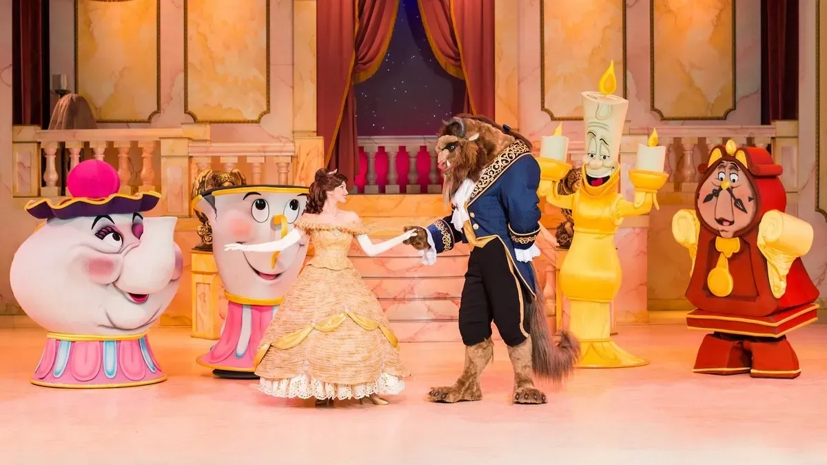 A colorful stage scene features characters from "Beauty and the Beast." Belle, in a golden gown, dances with the Beast, who is dressed in a blue and gold coat. They are surrounded by lively characters, including Mrs. Potts, Chip, Lumière, and Cogsworth, in a grand ballroom setting with ornate decor and warm lighting.