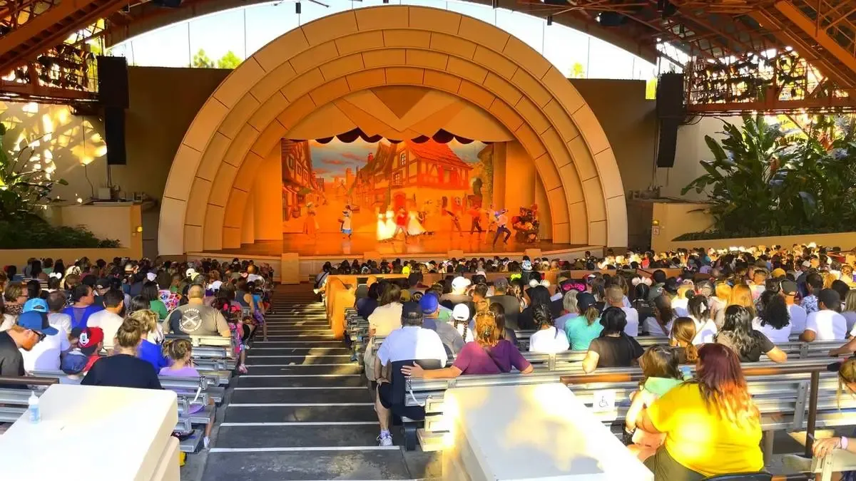 A large audience watches a stage performance under an open-air amphitheater with a distinctive arch design. The stage features a Beauty and the Beast show. The crowd is seated on tiered benches, enjoying the show.