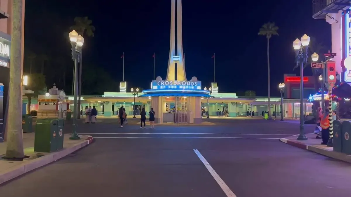 A nighttime view of a bright entrance area with an art deco design, featuring a tall spire and a sign reading "CROSSROADS." Several people are walking towards the entrance, and street lamps illuminate the scene.