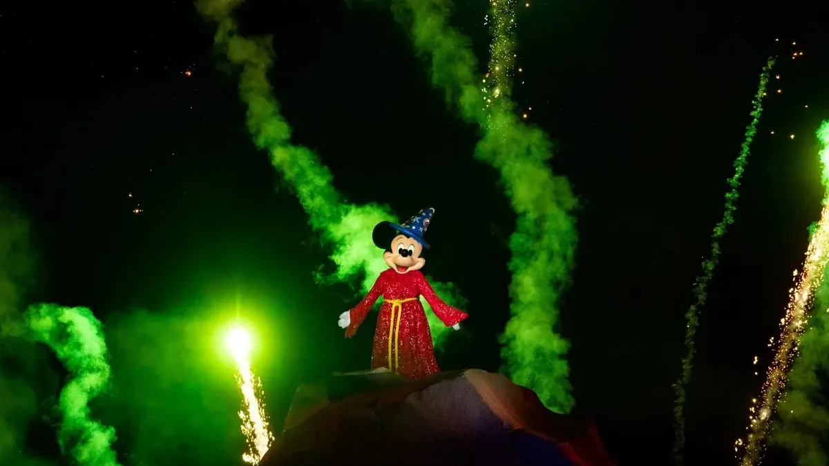 Mickey Mouse, dressed as a sorcerer in a red robe and blue hat, stands atop a rock as vibrant green fireworks light up the night sky behind him.