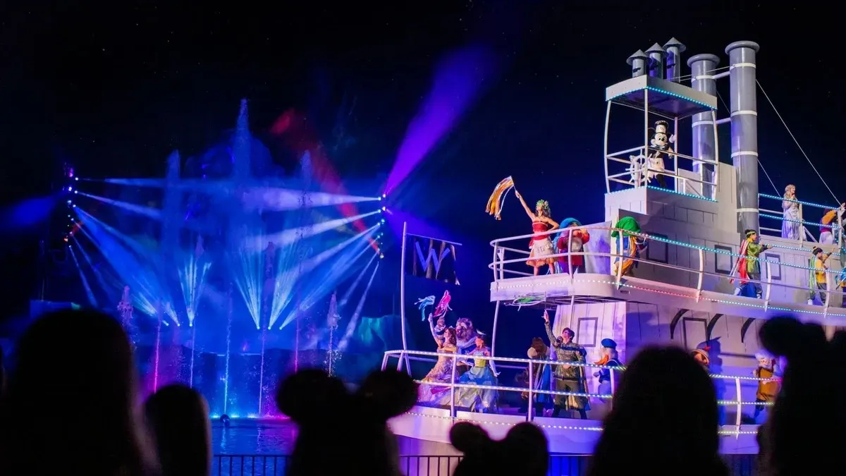 A nighttime performance features a large and illuminated riverboat with characters in colorful costumes dancing on deck. Bright lights and water fountains create a vibrant backdrop, as silhouettes of audience members with mouse ears watch in the foreground.