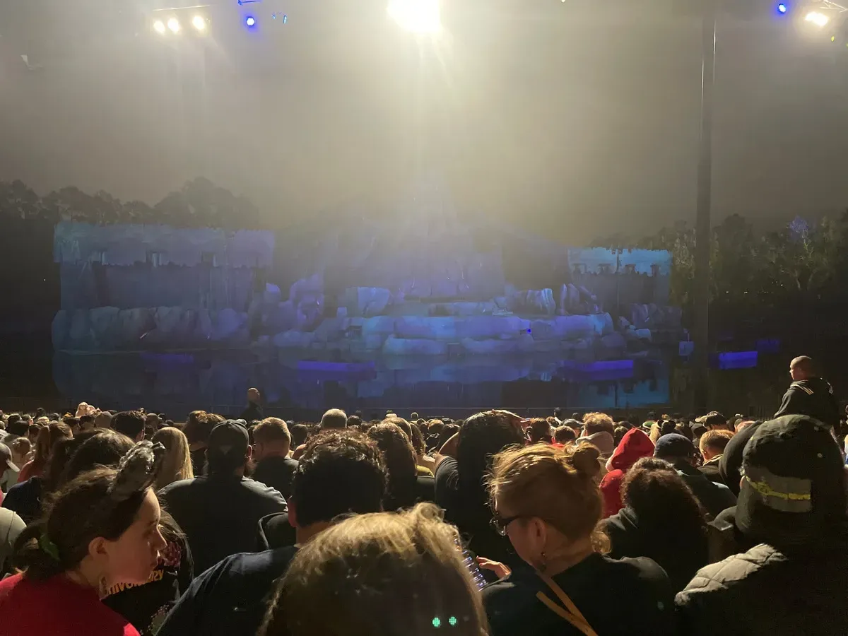 A crowd of people, many wearing raincoats and hoods, sits outdoors facing a stage with rocky scenery and blue lighting. It's nighttime, and beams of light shine through a cloudy sky, creating a dramatic atmosphere.