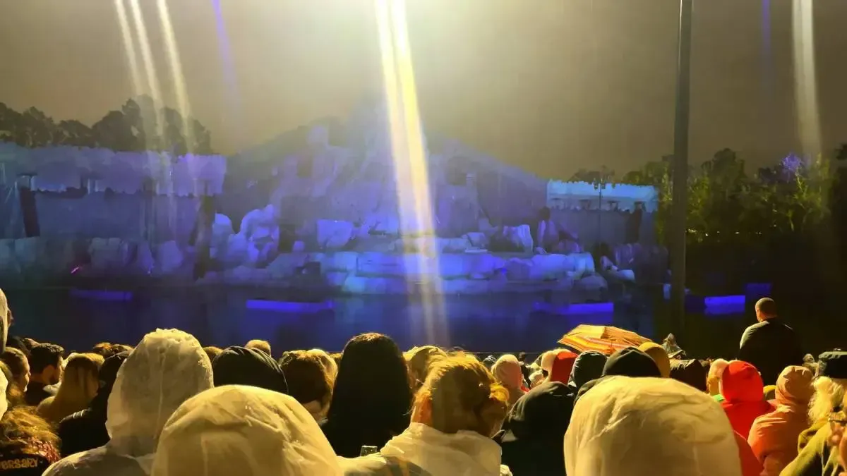 A large crowd gathers in an outdoor amphitheater at night, facing a stage designed to resemble a rocky, icy landscape. Bright lights illuminate the scene, casting a warm glow over the audience, who are eagerly awaiting a performance.