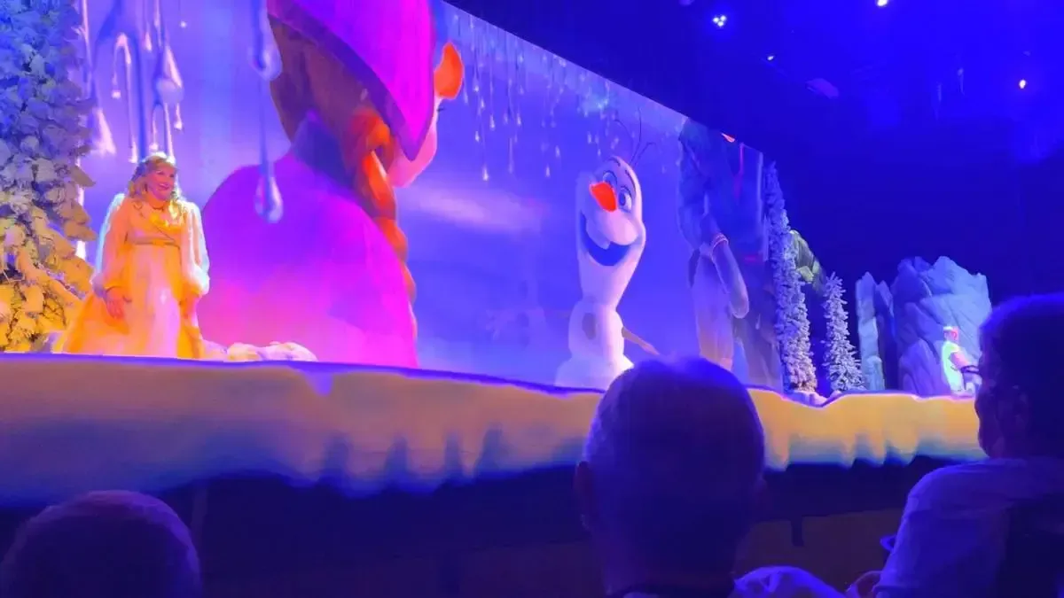 A Frozen stage performance features a person dressed in a white gown standing near a snowy tree, with a large screen displaying Anna and Olaf, under vibrant blue lighting. Audience members are visible in the foreground, watching the show.