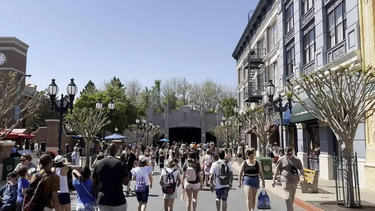 A bustling crowd of people strolls down a theme park street lined with ornate buildings and trees, under a clear blue sky.