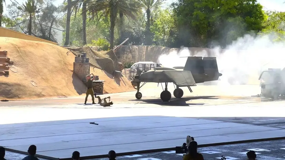 A dramatic action scene unfolds with a person in military attire dragging another person on the ground near a small aircraft. Smoke billows around the plane, adding intensity to the scene. The background features a sandy embankment and lush trees, creating a dynamic and engaging atmosphere.