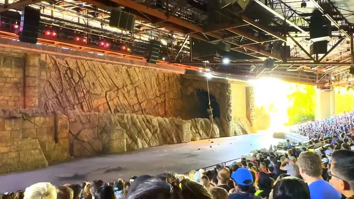 A large audience watches an indoor stage show featuring a rocky set with intricate stone textures and a person suspended by a rope. Bright lights illuminate the scene, while the ceiling is lined with technical equipment and lights.