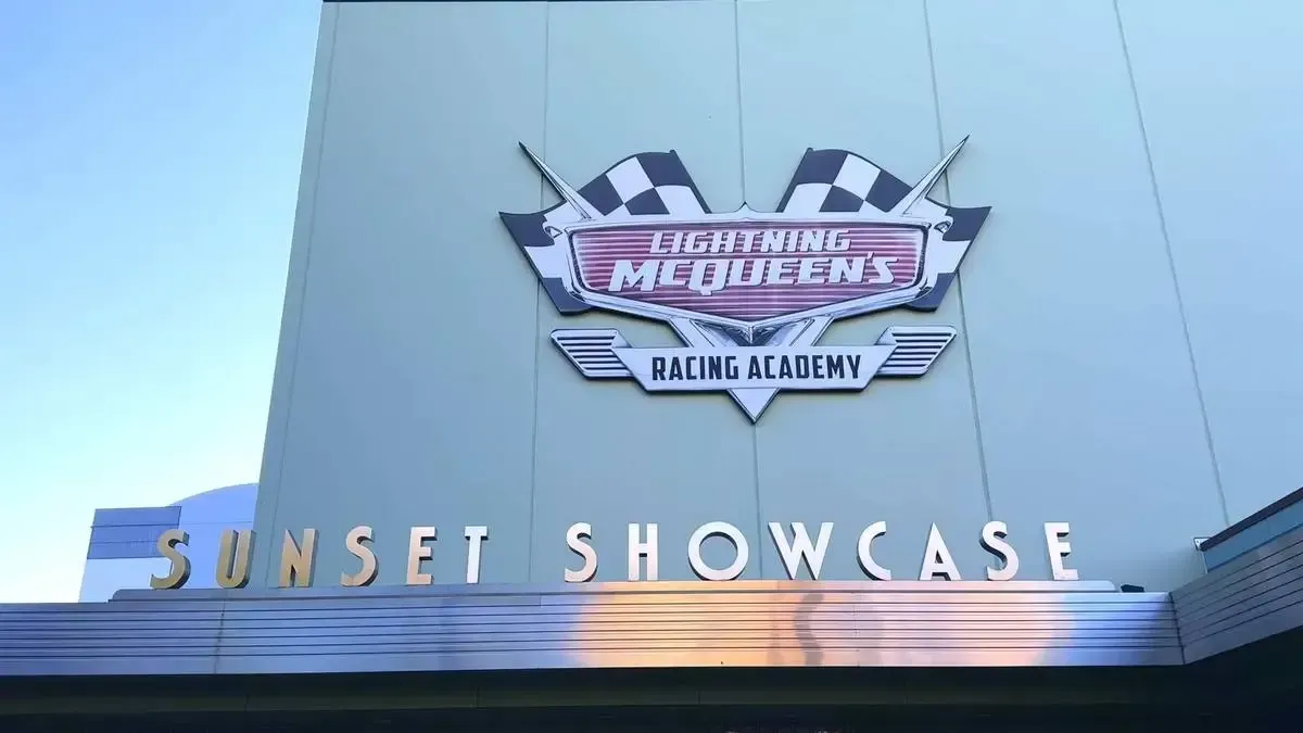 The image shows the entrance to Sunset Showcase featuring a sign for "Lightning McQueen's Racing Academy" with checkered flags, set against a light blue building under a clear sky.
