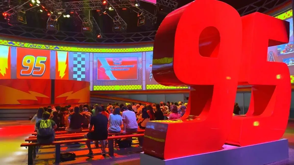 A large and red "95" sculpture dominates the foreground, while a crowd of people sit on benches in a colorful auditorium. The walls display dynamic graphics featuring the number 95 and racing motifs, creating an energetic atmosphere.