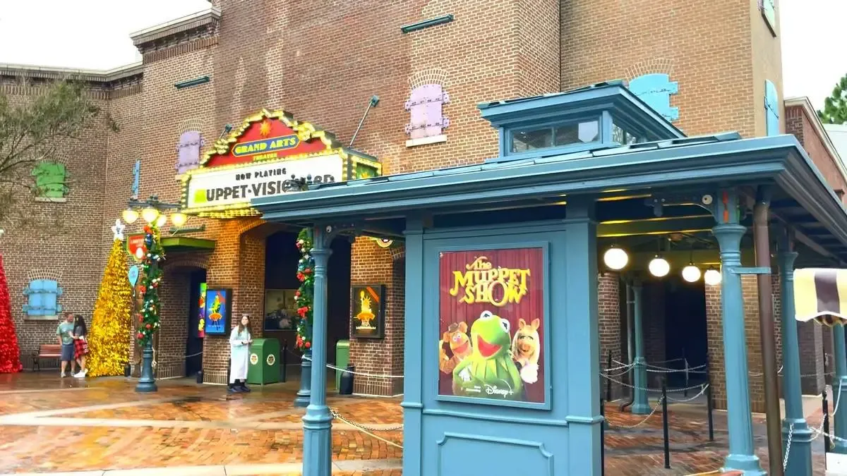 The entrance to a theater features a colorful sign for "Muppet-Vision 3D" on a brick building, adorned with festive decorations and a poster for "The Muppet Show" with Kermit the Frog and friends.