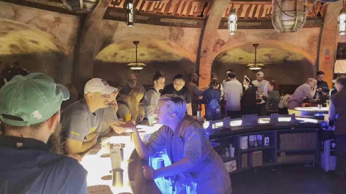 A bar scene with people gathered around a curved, illuminated counter. The setting features rustic, arched walls and warm lighting. Groups of people are engaged in conversation, with drinks in hand, creating a bustling and social atmosphere.