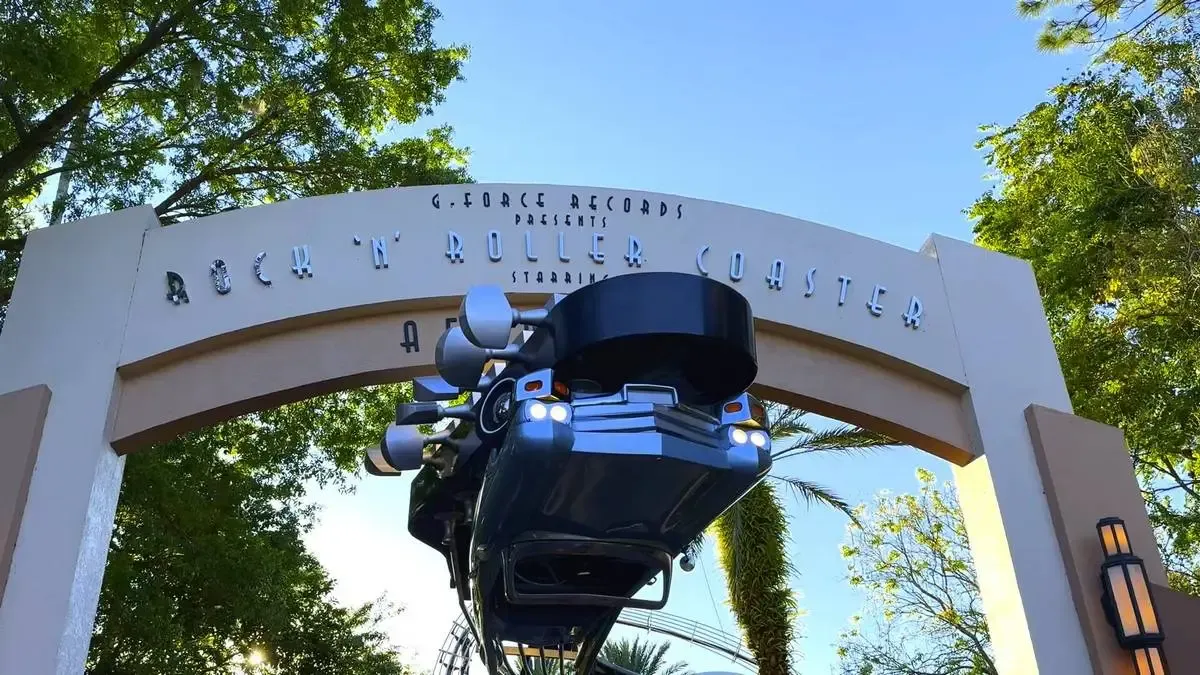 The entrance to the Rock 'n' Roller Coaster features a large archway with the ride's name and a sleek black car suspended above, set against a backdrop of green trees and a clear blue sky.