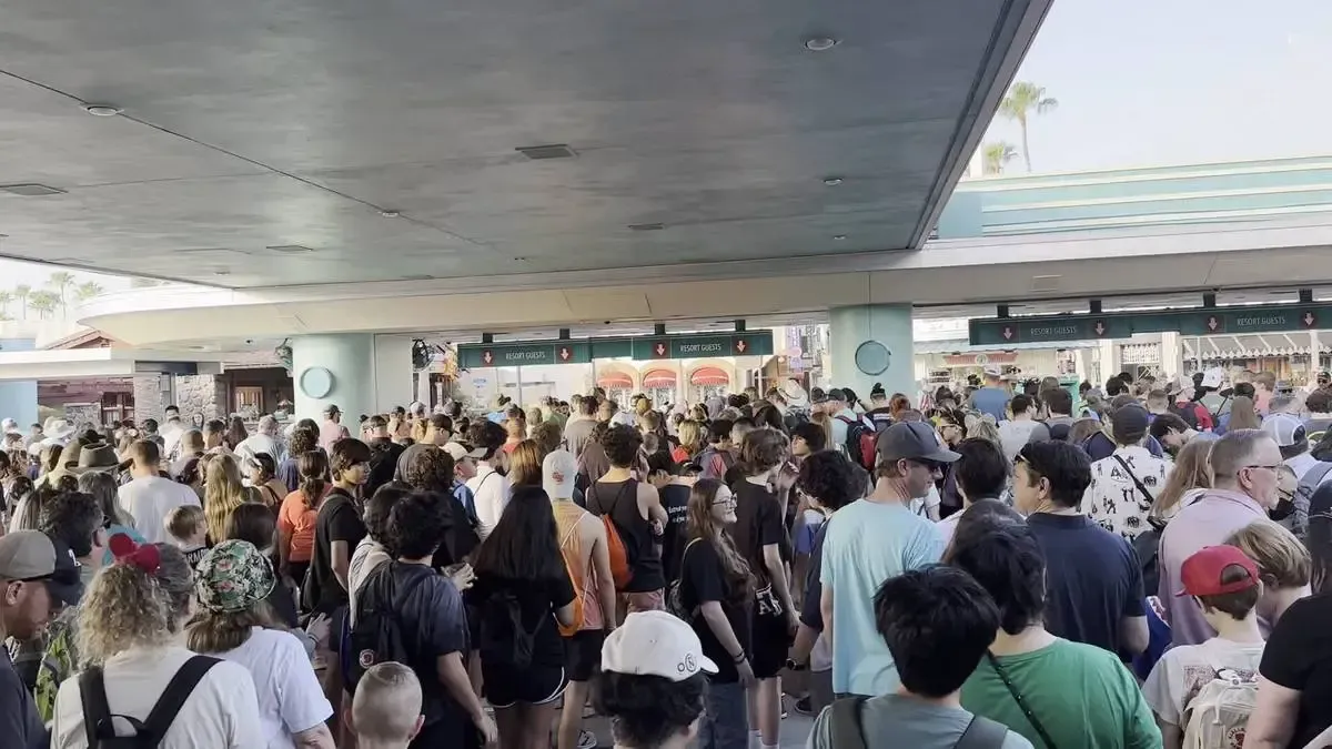 A large crowd of people gathers under a covered area, waiting in line at Hollywood Studios entrance. The scene is bustling with individuals wearing casual clothes, hats, and backpacks, suggesting a busy day at the theme park.