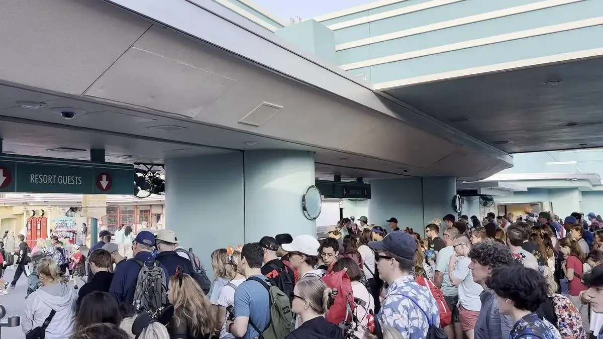 A large crowd of people waits in line under a covered area marked "Resort Guests," with many wearing backpacks and casual clothing. The setting appears to be an entrance to a theme park or resort, with blue-green walls and overhead signage.
