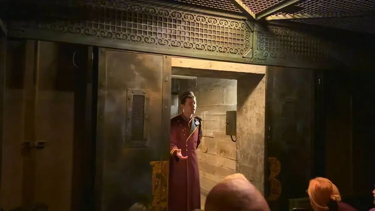 A Tower of Terror cast member in a maroon uniform stands in a industrial-style room with metal walls and intricate latticework. They appear to be speaking to a group of people entering the ride. The setting has an eerie, vintage atmosphere.