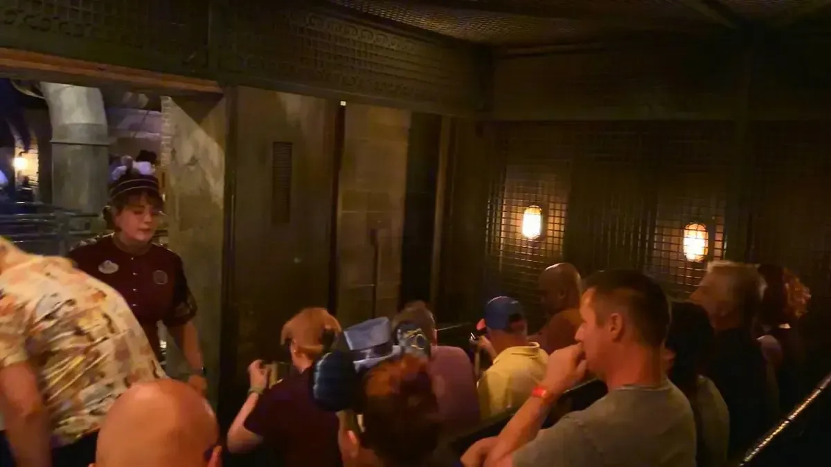 A group of people waits in line inside a dimly lit, themed attraction area, with a staff member in a uniform guiding them. The setting features textured walls and warm lighting, creating an immersive atmosphere.