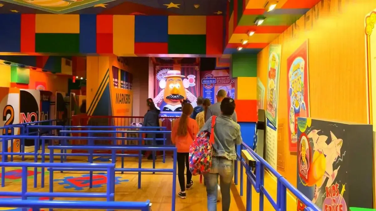 People walk through a colorful, toy-themed indoor space with vibrant walls and a large Mr. Potato Head figure in the background. Blue railings guide visitors, and playful posters decorate the walls.