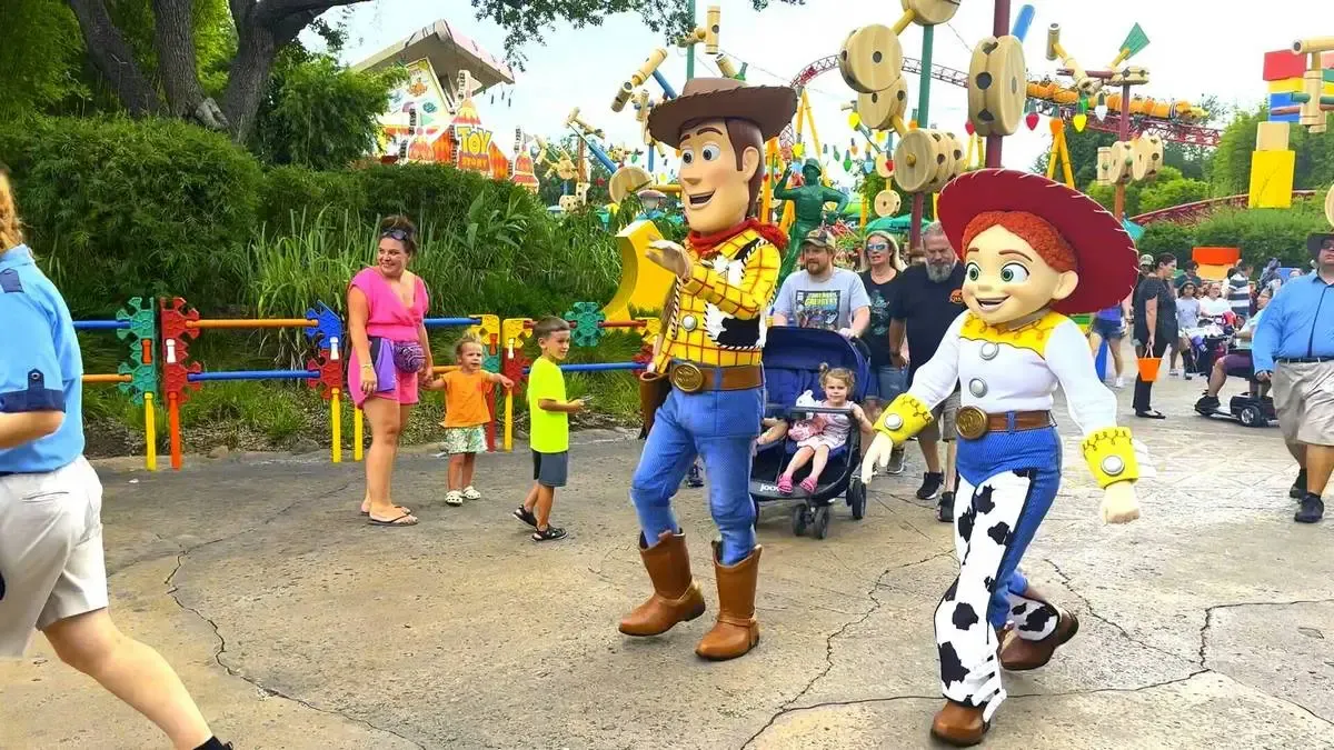 Woody and Jessie from Toy Story walk cheerfully through a theme park, surrounded by excited visitors and colorful decorations, while children watch in delight.