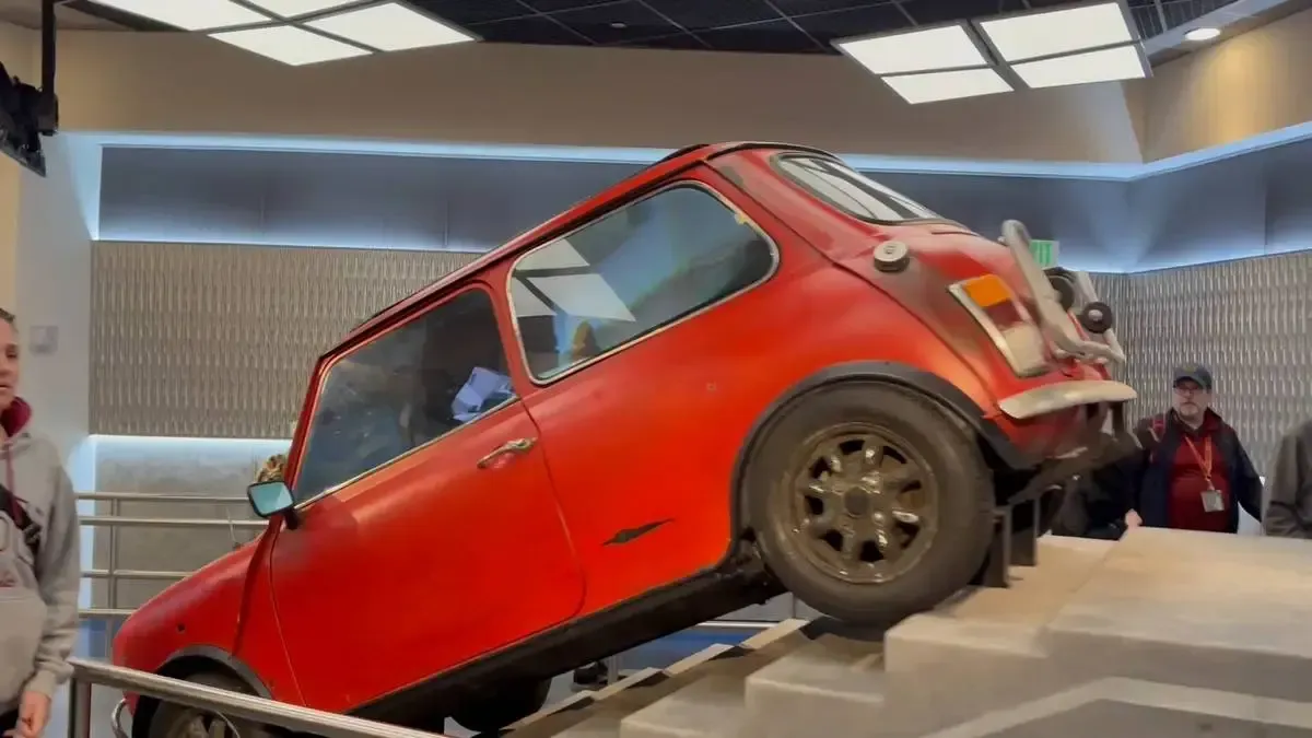 A vintage red Mini Cooper is displayed indoors, angled upwards on a set of concrete steps. The car's worn exterior contrasts with the modern interior setting, and several people stand nearby, observing the exhibit.