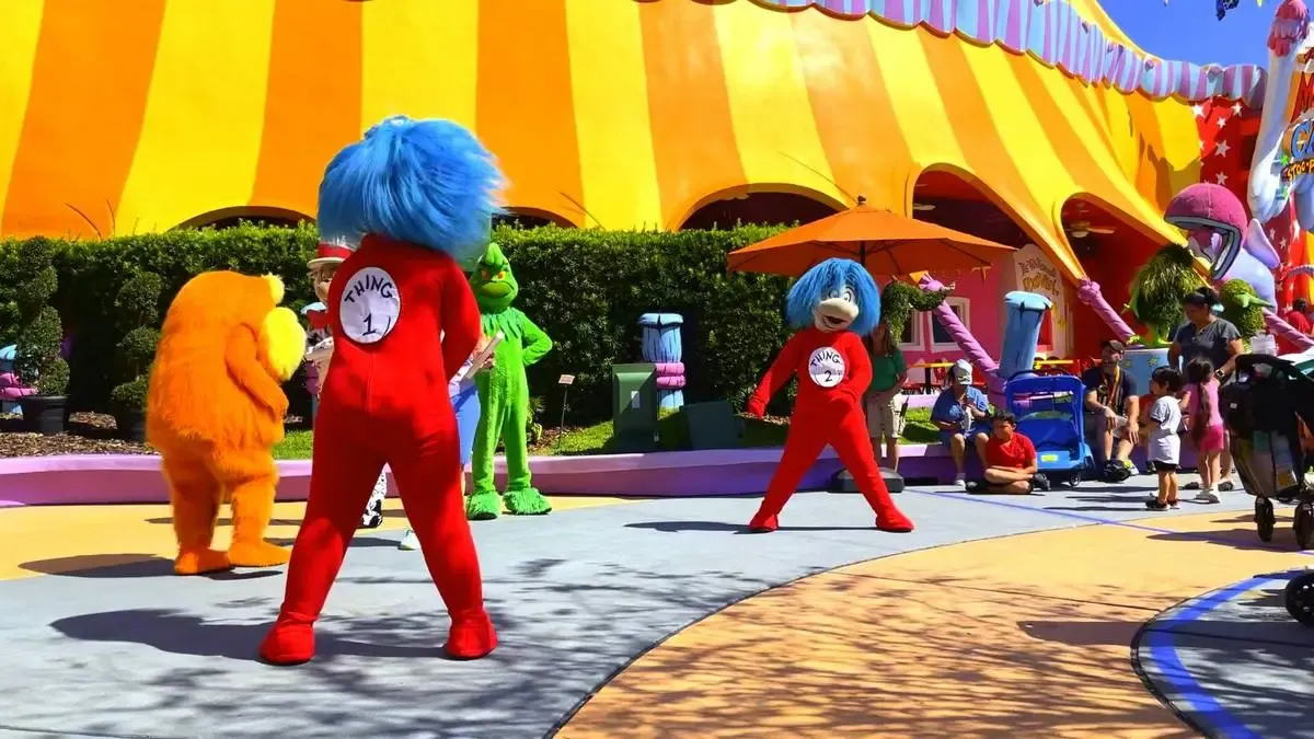 Costumed characters from Dr. Seuss stories, including Thing 1 and Thing 2, dance in front of a vibrant yellow and orange tent at a theme park. The scene is lively, with families and children watching nearby.