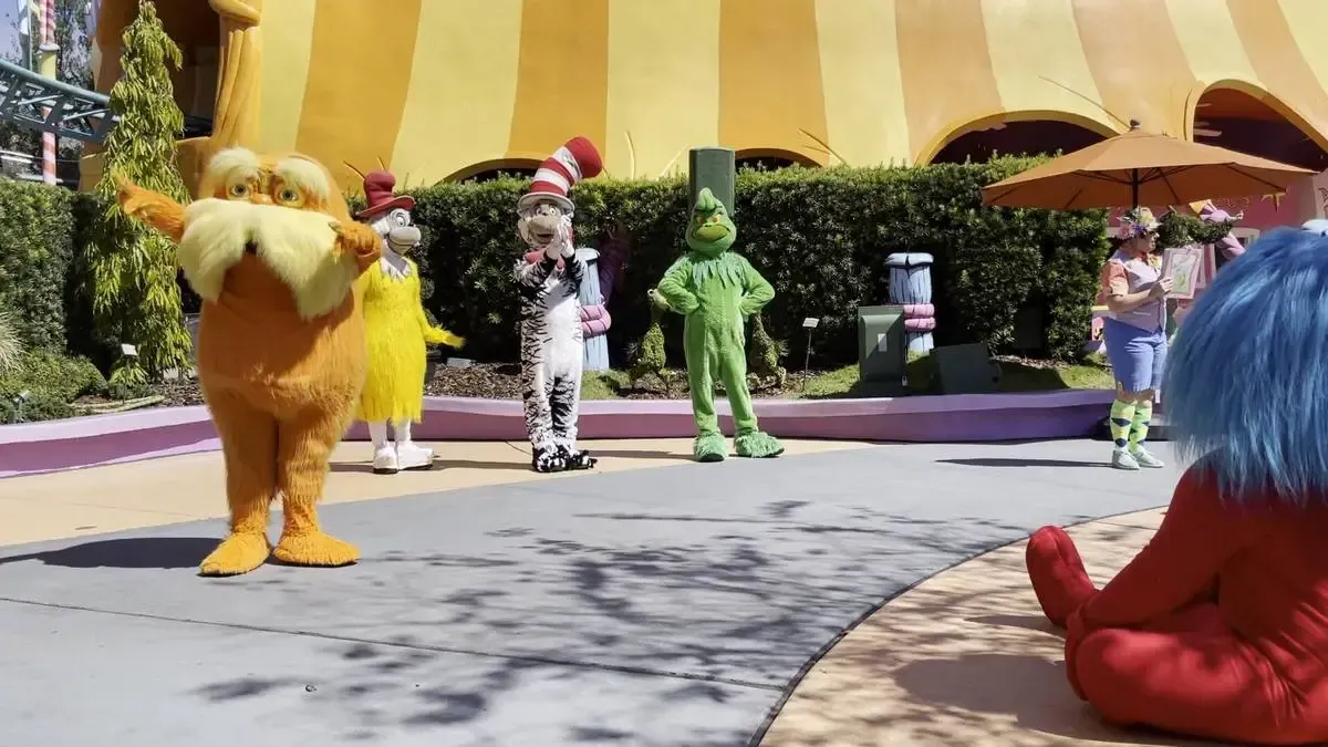 Costumed characters, including an orange creature with a large mustache, a tall figure in a striped hat, and a green figure, entertain visitors in a theme park setting with vibrant yellow and orange structures and lush greenery. A person in a colorful outfit stands nearby under an umbrella, while a seated figure with blue hair watches the scene.