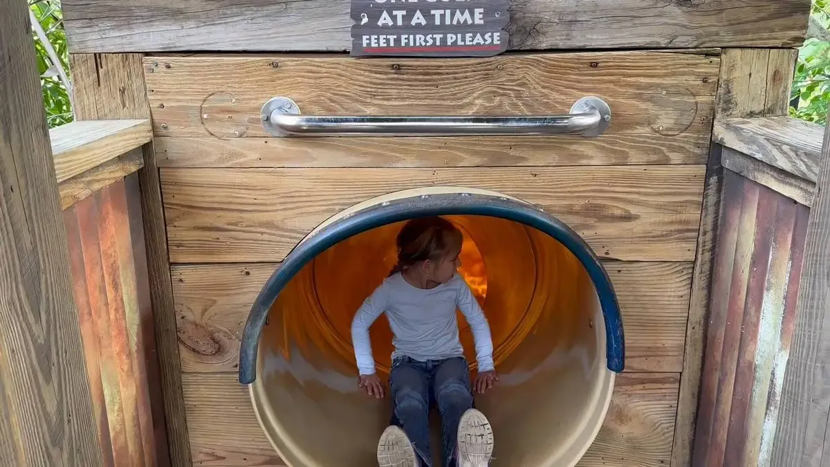 A child wearing a white long-sleeve shirt and jeans sits at the entrance of a wooden slide, preparing to go down feet first. The slide is part of a rustic play structure, and a sign above reads "One at a time, feet first please.".