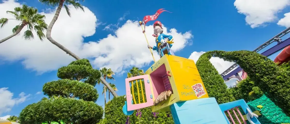 A whimsical statue of a character holding a red flag stands atop a colorful, yellow and pink box labeled "Tiger" in a lush, green theme park setting. The sky is bright blue with fluffy white clouds, and palm trees and manicured hedges surround the scene.