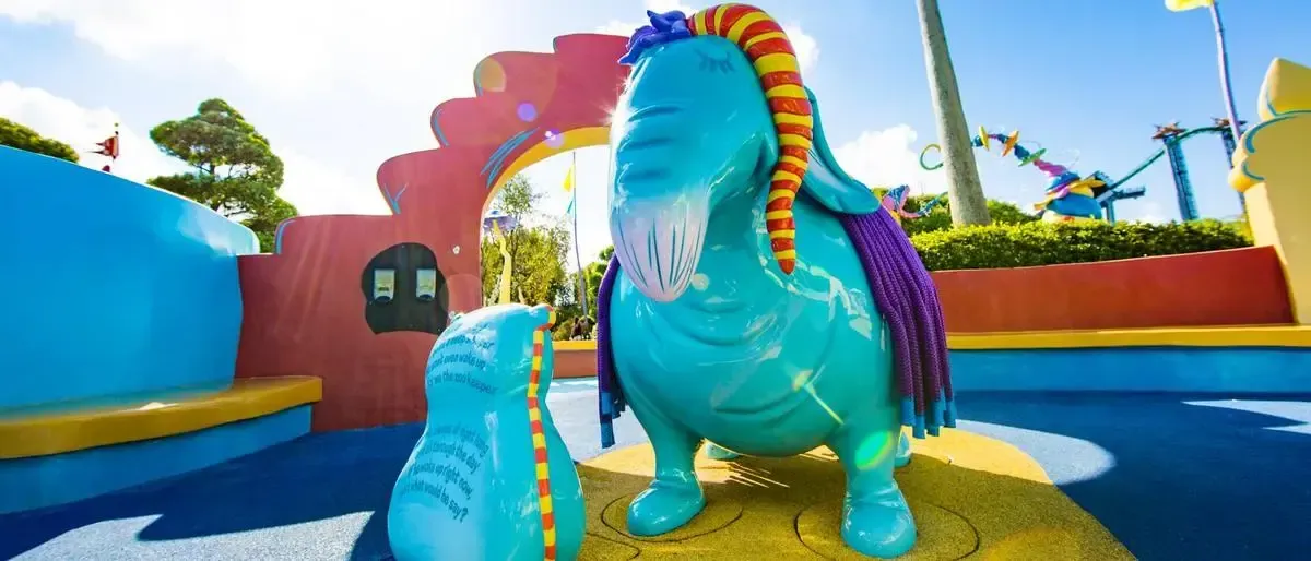 A whimsical blue statue of a creature with a colorful striped trunk and mane stands in a vibrant theme park setting, surrounded by playful, curved structures and bright colors.
