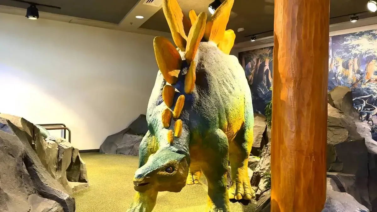A colorful Stegosaurus model stands in a museum exhibit, surrounded by artificial rocks and a large wooden post. The dinosaur features vibrant shades of green and yellow, with distinctive orange plates along its back, under bright indoor lighting.