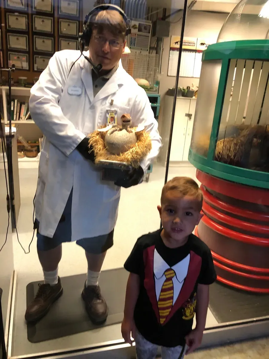 A man in a lab coat, wearing a headset, stands behind glass holding a nest with a hatching dinosaur egg. A young boy in a novelty shirt stands in front, looking at the camera. The setting resembles a science exhibit with equipment and certificates visible in the background.