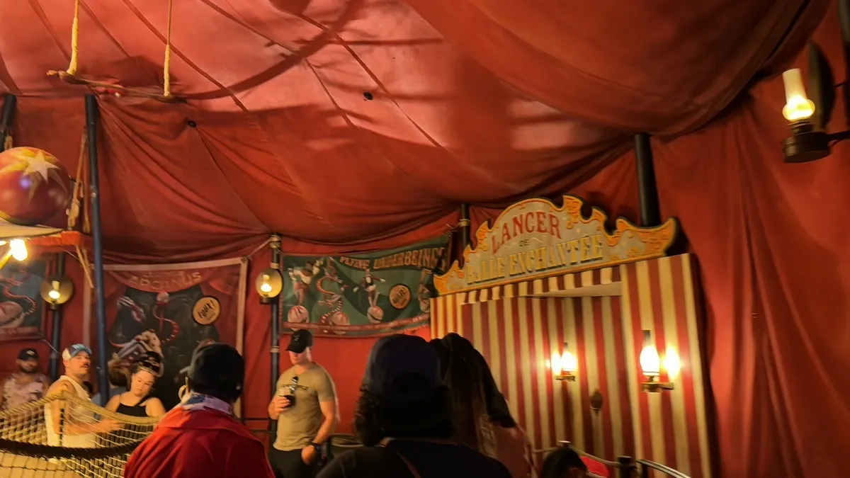 A group of people stands inside a circus-themed room with red draped ceilings and striped walls. Vintage-style posters and signs, including one that reads "Lancer de Balles Enchantées," decorate the space. Soft, warm lighting from antique-style lamps enhances the nostalgic atmosphere.