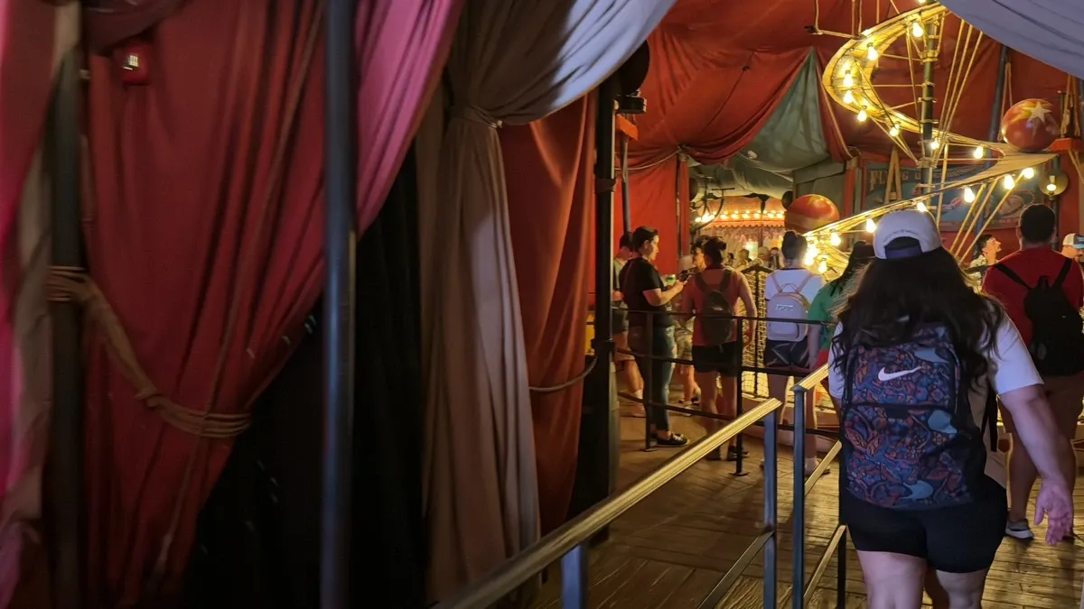 A group of people walk through a dimly lit, tent-like hallway adorned with colorful drapes and a spiral of glowing lights. The atmosphere is lively, with a woman in the foreground wearing a patterned backpack and a white cap.