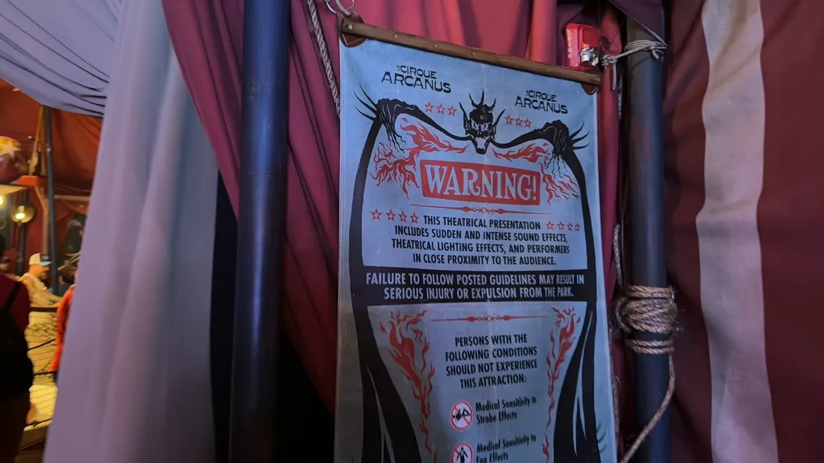 A warning sign for Cirque Arcanus is displayed inside a tent, cautioning about sudden sound effects and theatrical lighting. The sign advises individuals with certain medical sensitivities to avoid the attraction.