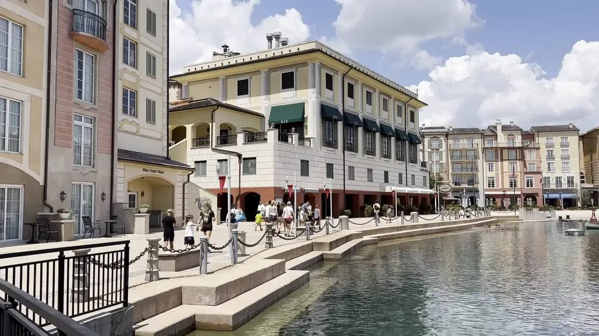 A charming waterfront scene features colorful buildings with balconies and awnings, reminiscent of an Italian village. People stroll along the promenade beside a calm, reflective body of water under a partly cloudy sky.
