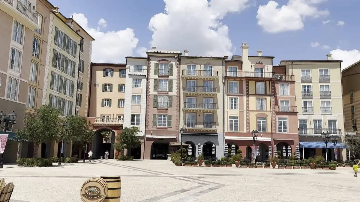 A charming plaza is surrounded by colorful buildings with classic European architecture. The facades feature balconies and shuttered windows. Potted plants and trees line the open, sunlit square, and a few people stroll under a bright blue sky with fluffy clouds.