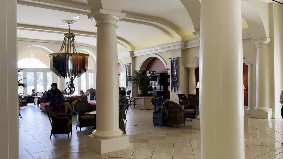 A spacious hotel lobby features elegant archways and tall columns. Comfortable seating areas with wicker furniture are arranged throughout, and a large, ornate chandelier hangs from the ceiling.