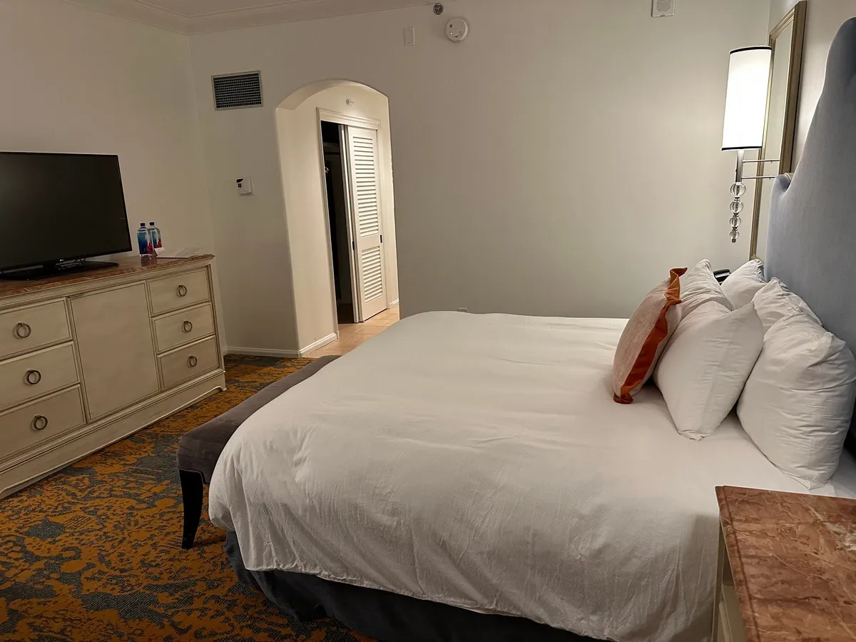 A cozy hotel room features a bed with multiple pillows. A dresser with a flat-screen TV and two water bottles sits across from the bed, and a patterned carpet adds warmth to the room.