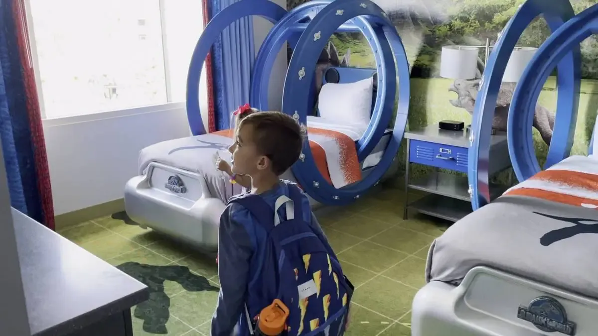 A child with a backpack inside a themed hotel room featuring two unique, circular blue beds. The room is decorated with dinosaur imagery, including a mural of a dinosaur and beds adorned with orange and gray bedding.