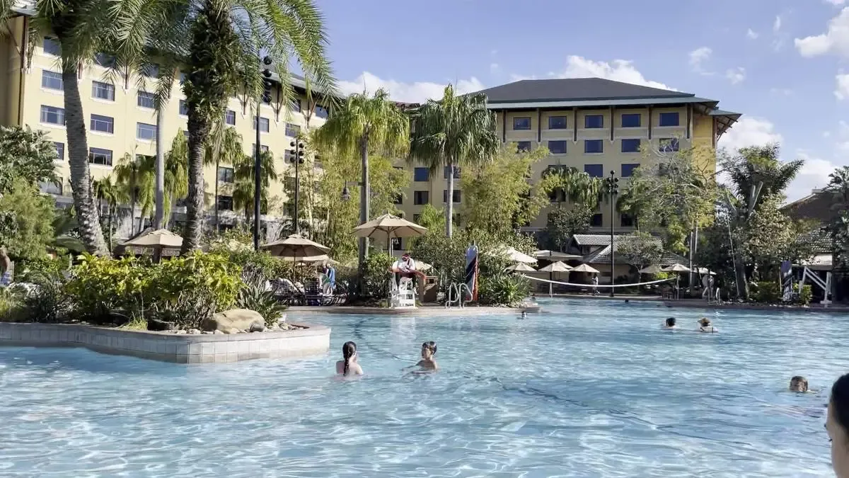 People enjoy swimming in a large, sunlit pool surrounded by lush palm trees and tropical plants, with a multi-story hotel building in the background under a clear blue sky.