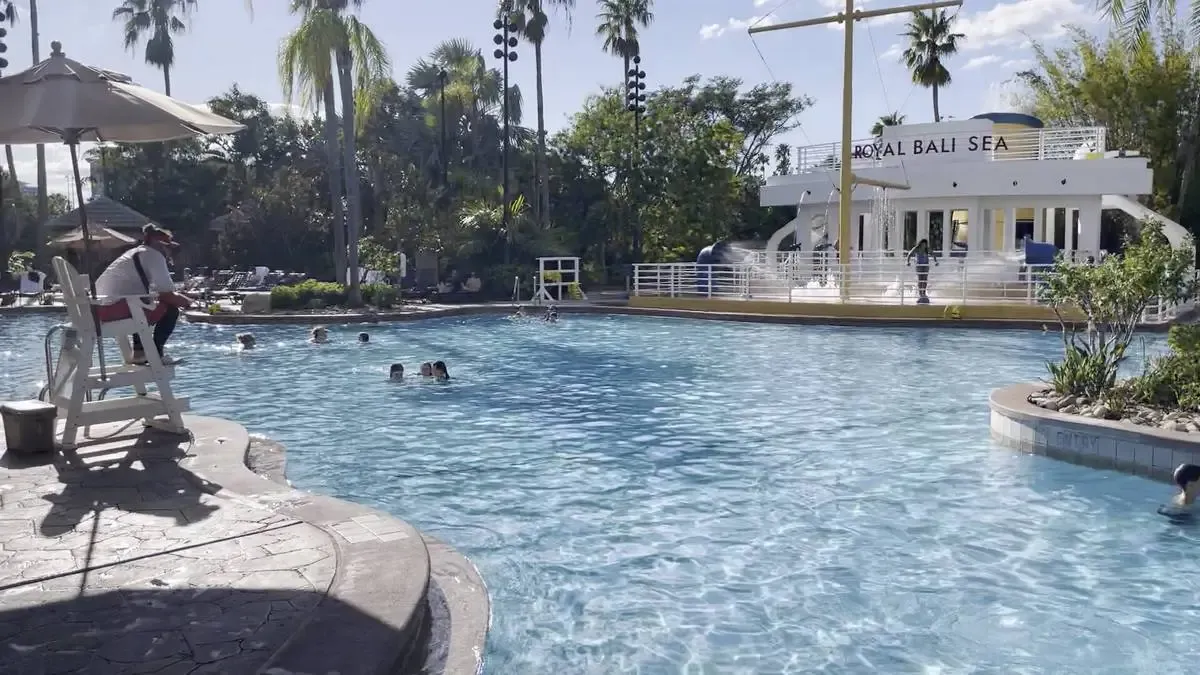 A sunny pool scene with a lifeguard sitting under an umbrella on a raised chair, overseeing swimmers enjoying the water. In the background, a structure labeled "Royal Bali Sea" is visible, surrounded by lush palm trees and greenery.