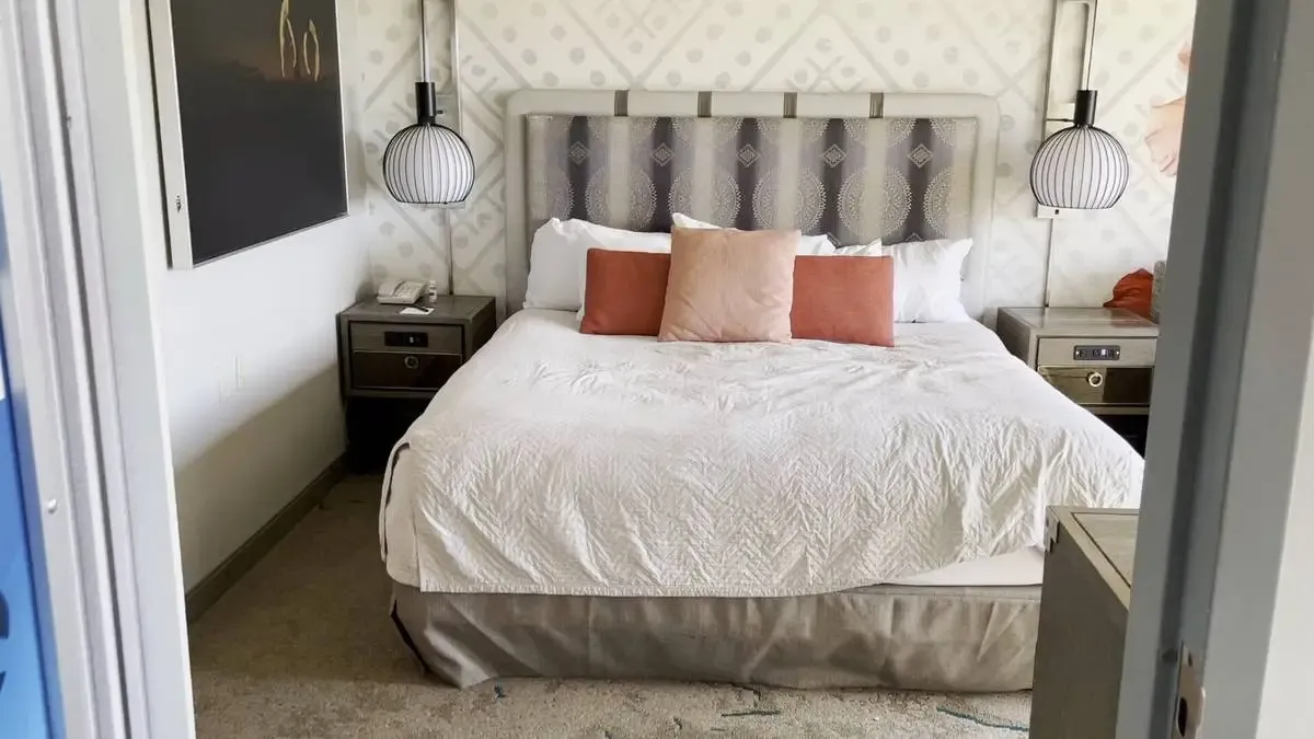 A cozy bedroom features a bed with a white comforter and three decorative pillows in peach and orange hues. Two modern pendant lights hang above matching bedside tables, and a patterned wallpaper adds a stylish touch to the space.